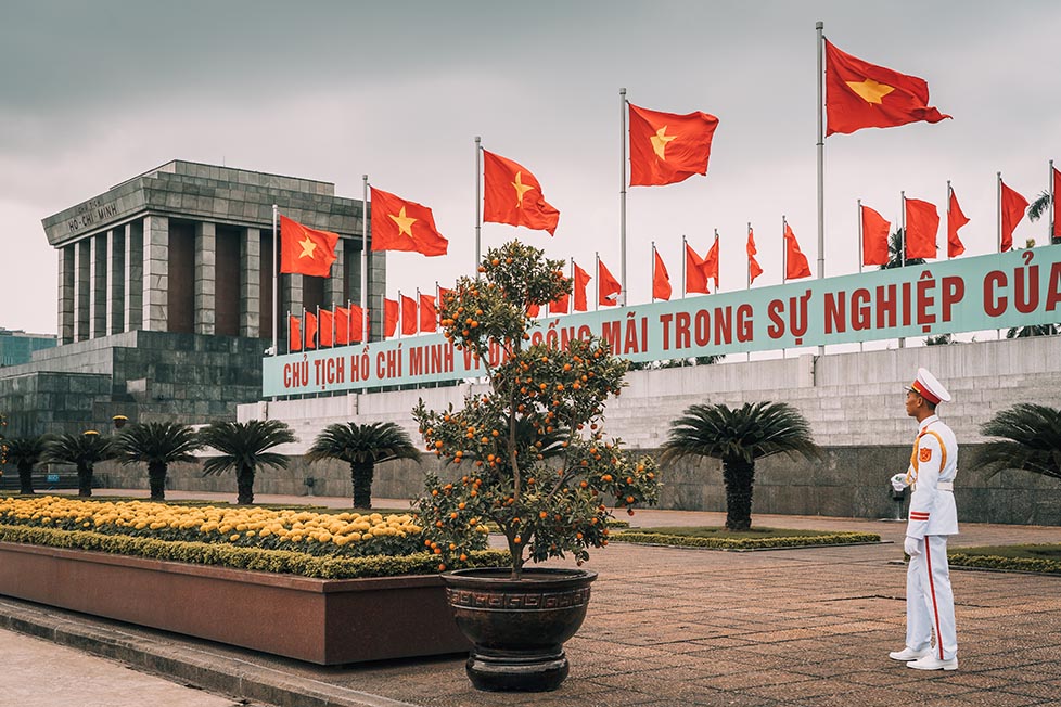 A soldier in a white uniform is standing next to a row of Vietnamese flags outside the Ho Chi Minh Mausoleum in Hanoi, Vietnam