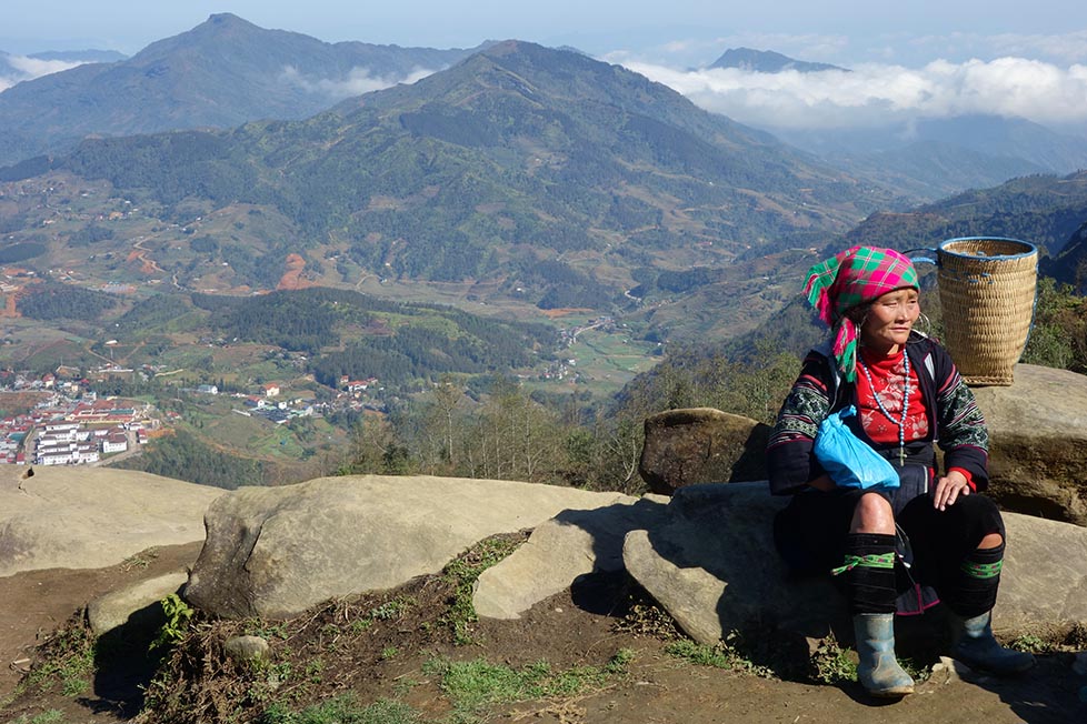 A H'mong tribal woman wearing traditional clothing sitting on a rock on a hillside with a valley below and massive mountains with low cloud hugging them behind her in Sapa Vietnam