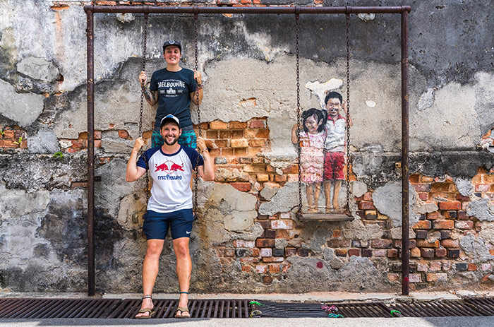 Nic and Shorty on a swing on the interactive street art of Georgetown, Penang in Malaysia with a crumbly wall behind them