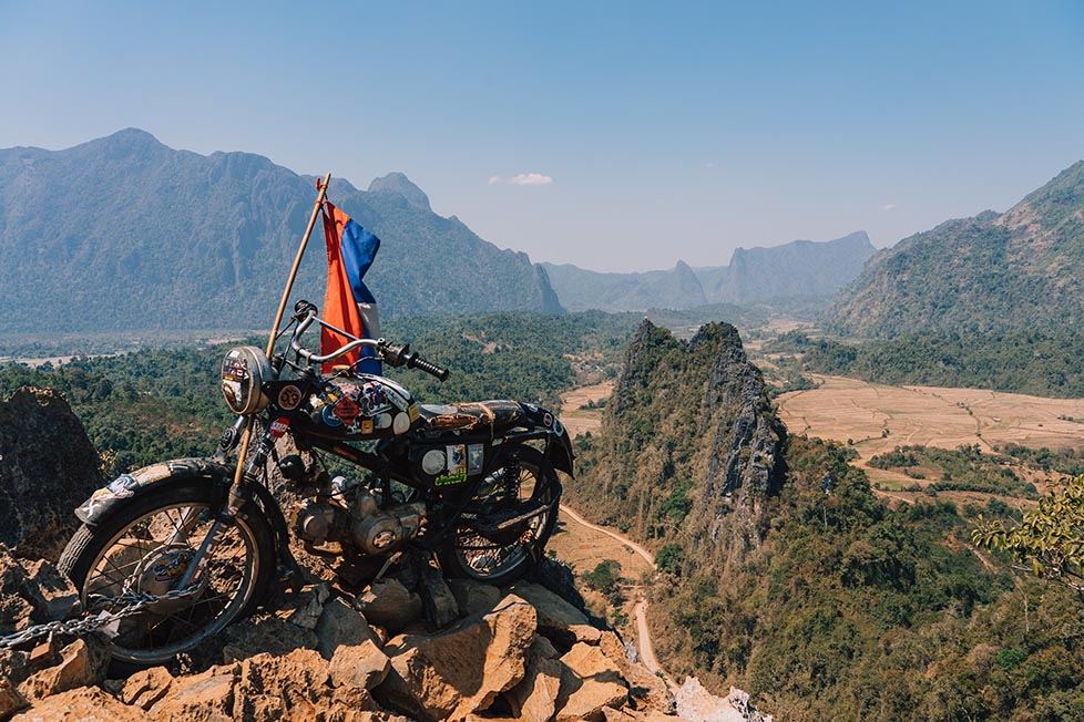 An old motorbike with a Laotian flag on it at the top of a mountain with other mountains in the distance in the town of Vang Vieng, Laos