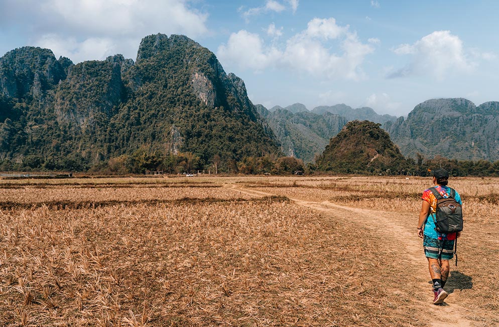 Nic walking across a farmers field in Vang Vieng, Laos toward the giant jungle covered karst mountains, in Laos