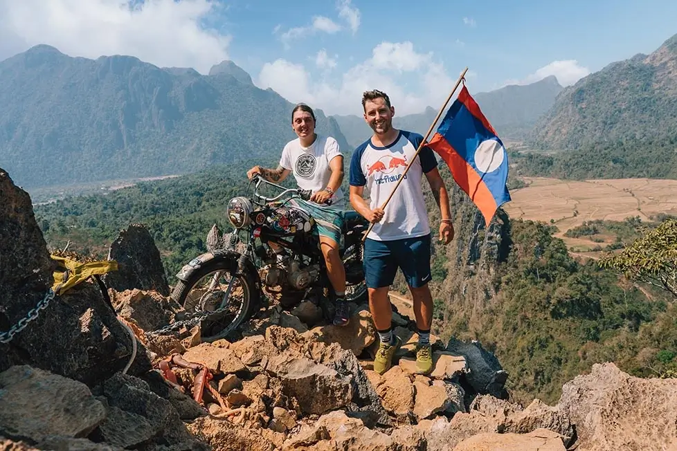 Nic and Shorty of The Roaming Renegades standing on a summit in Laos and smiling broadly while holding a Laotian flag.
